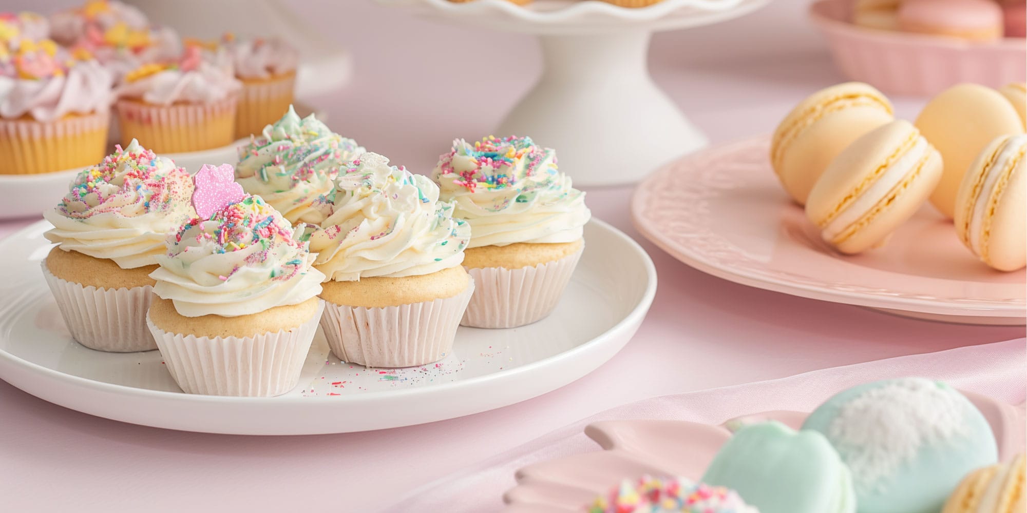 Carefully crafted vanilla dessert cupcakes with delicate frosting and a scattering of colorful sprinkles, presented alongside elegant, pale yellow macarons on a pink platter. A perfect little break.