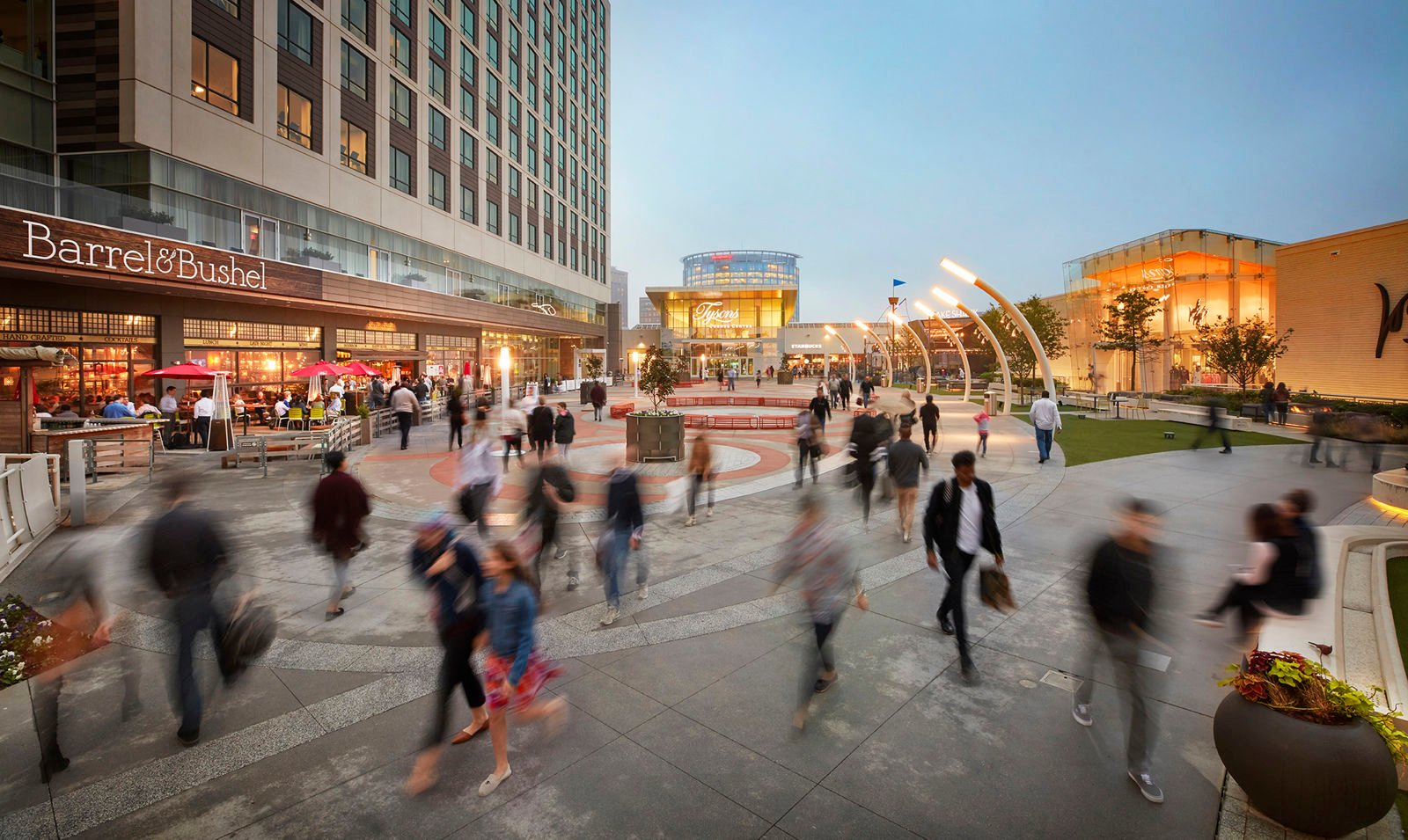 An evening shot of the outdoor plaza at Tysons Corner Mall, showing a modern urban square with blurred figures of people walking. The area is bordered by restaurants with outdoor seating, and the warm lights from the buildings create a welcoming, vibrant atmosphere.