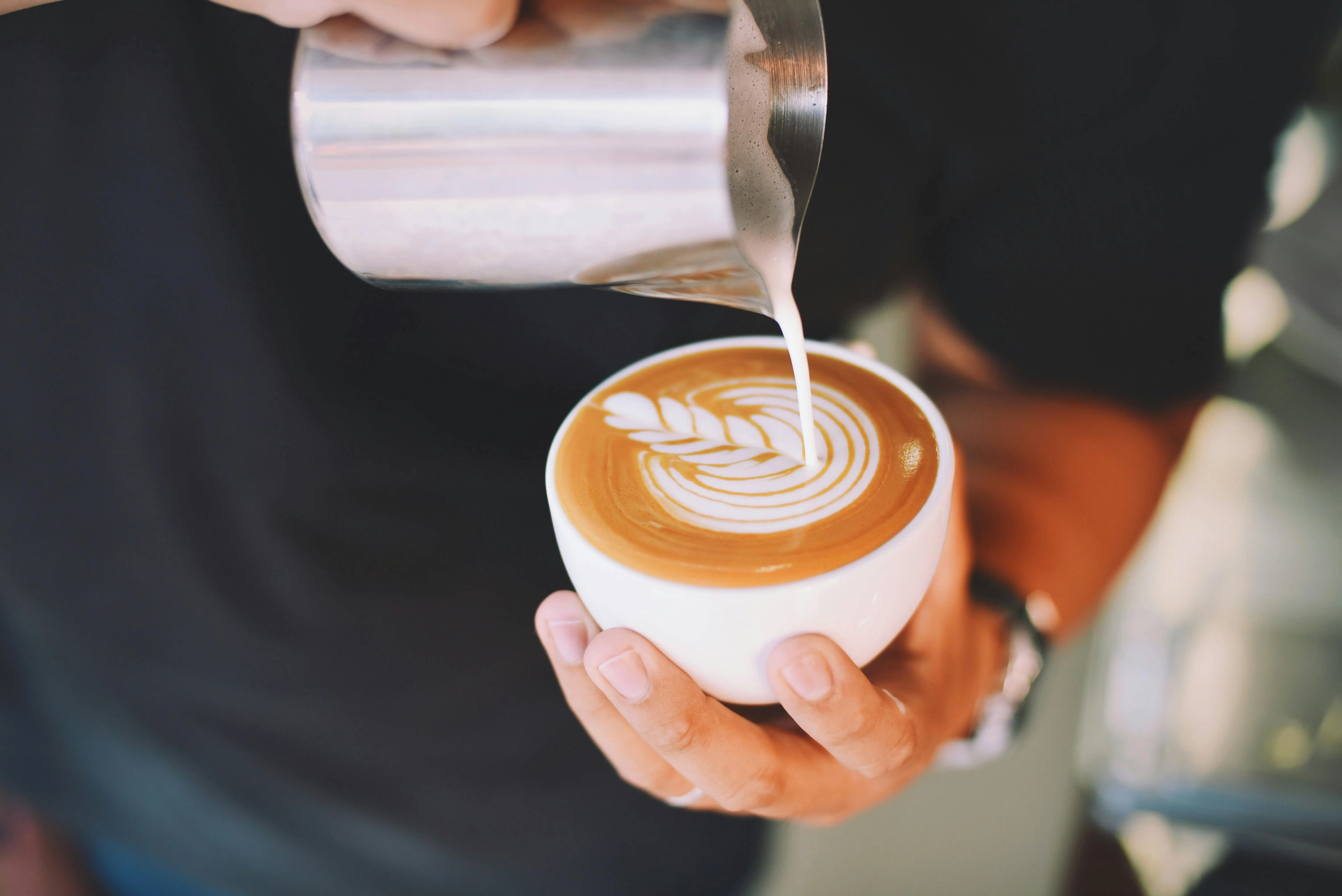 A close-up image captures a barista pouring steamed milk from a metal pitcher into a white cup, creating detailed latte art in the pattern of a fern or leaf. The cup contains a rich, dark base, suggesting a beverage made with fresh espresso coffee shots, and the final creation is a beautiful, creamy coffee drink.