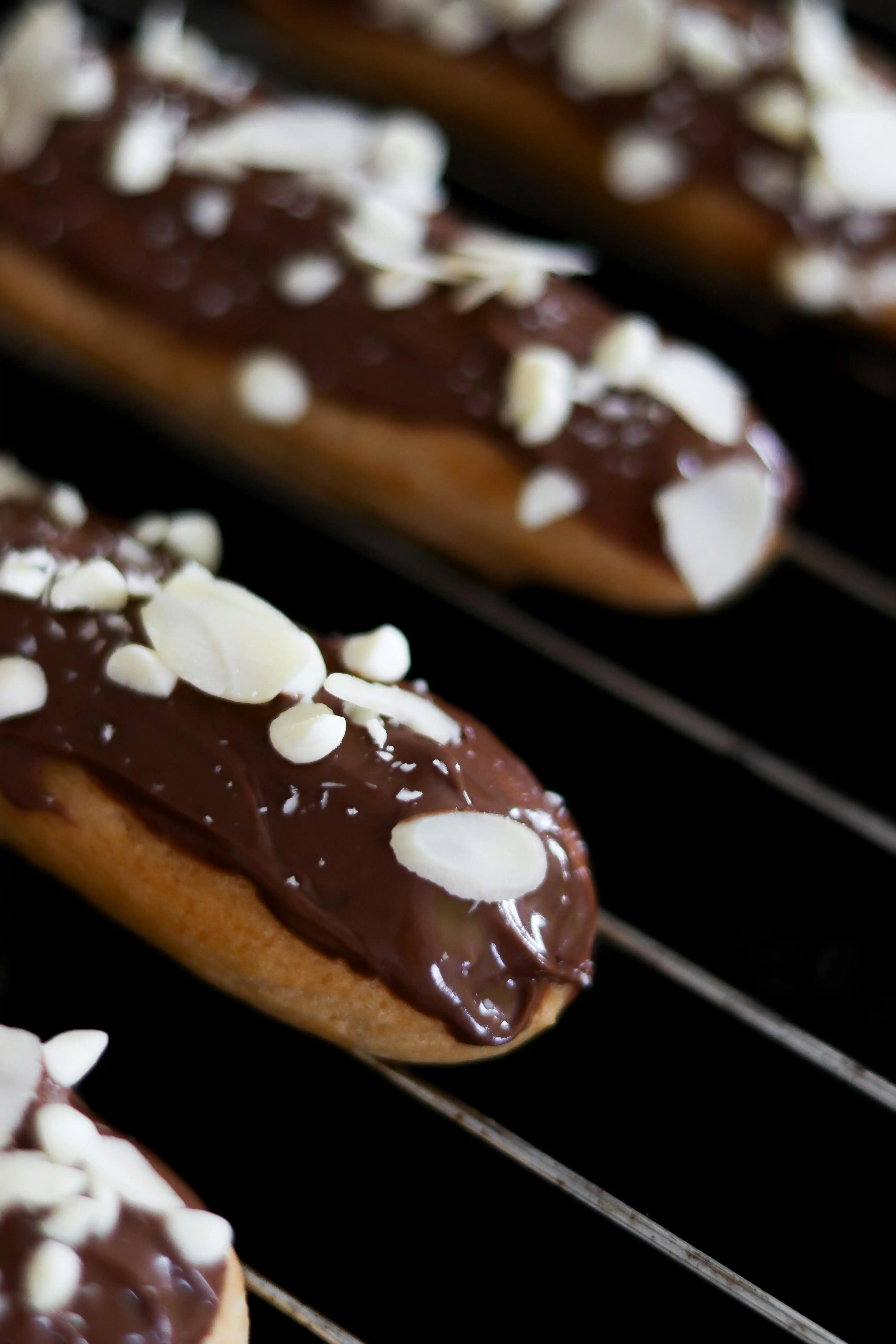 A close-up photograph displays several chocolate eclairs resting on a wire cooling rack. Each chocolate eclair is coated in a glossy dark chocolate glaze and liberally sprinkled with sliced almonds. The eclair in the foreground is in sharp focus, while the others become progressively blurred in the background.