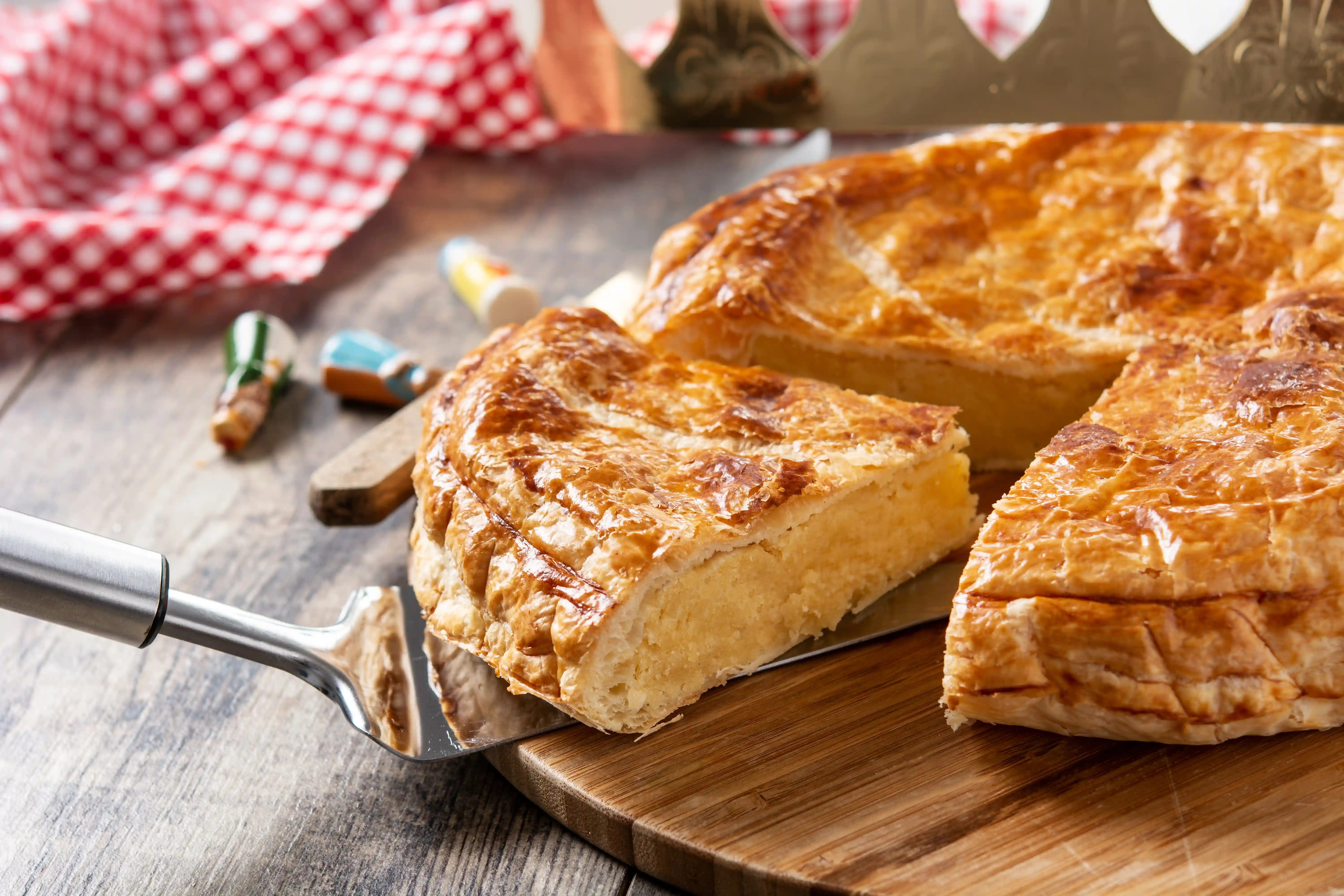 A golden-brown galette des rois sits on a wooden board, with one slice lifted away to reveal its dense frangipane filling. The flaky puff pastry crust features a traditional scored pattern and is accompanied by a golden paper crown and colorful ceramic charms. A rustic setting with a red-and-white checkered cloth completes the festive scene of this French Epiphany tradition.