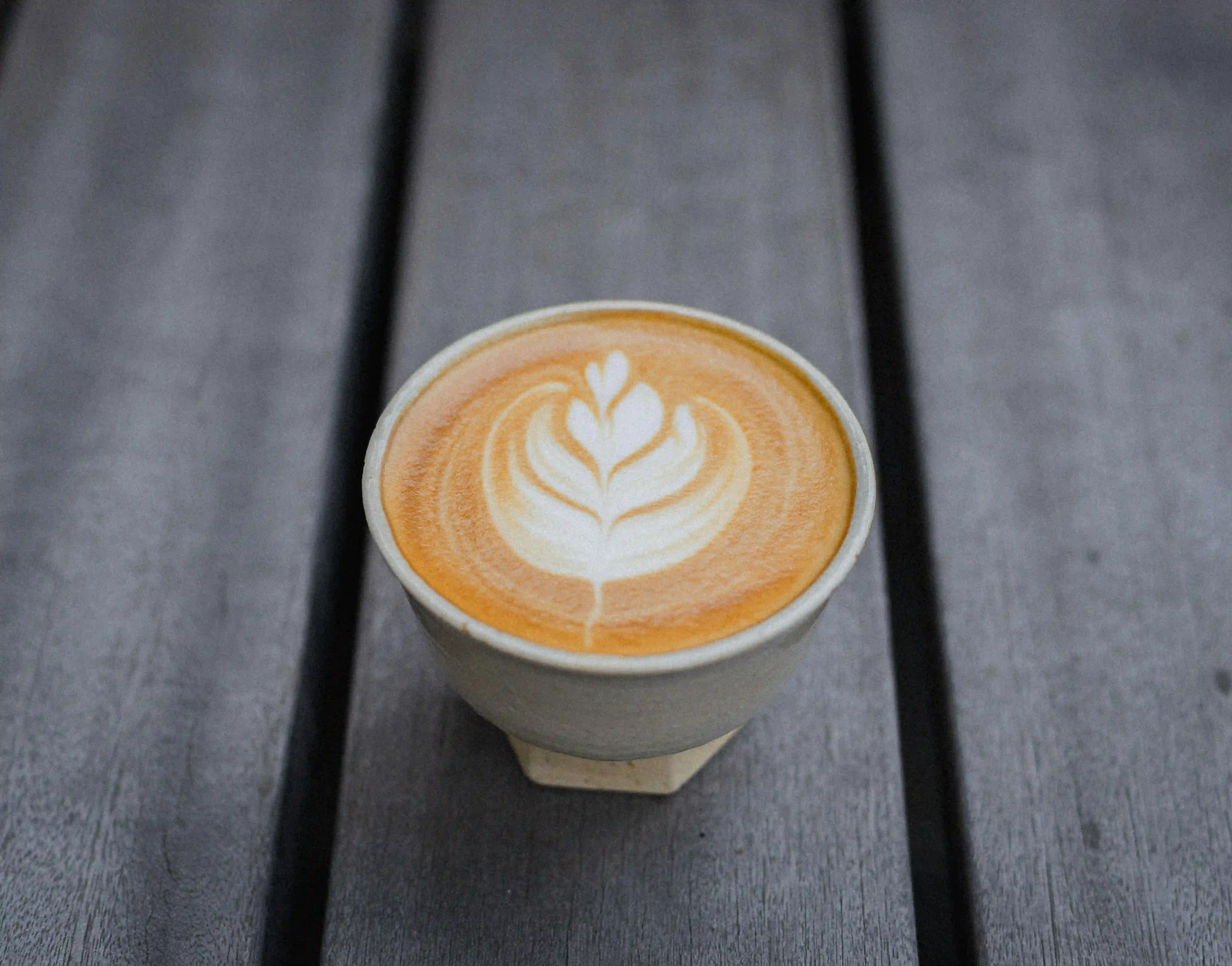 A small ceramic cup of latte sits on a gray wooden table, topped with delicate leaf-shaped latte art. The warm espresso crema frames the white foam pattern in the center. Perfect for anyone searching “coffee near me” and craving a calm, cozy moment.