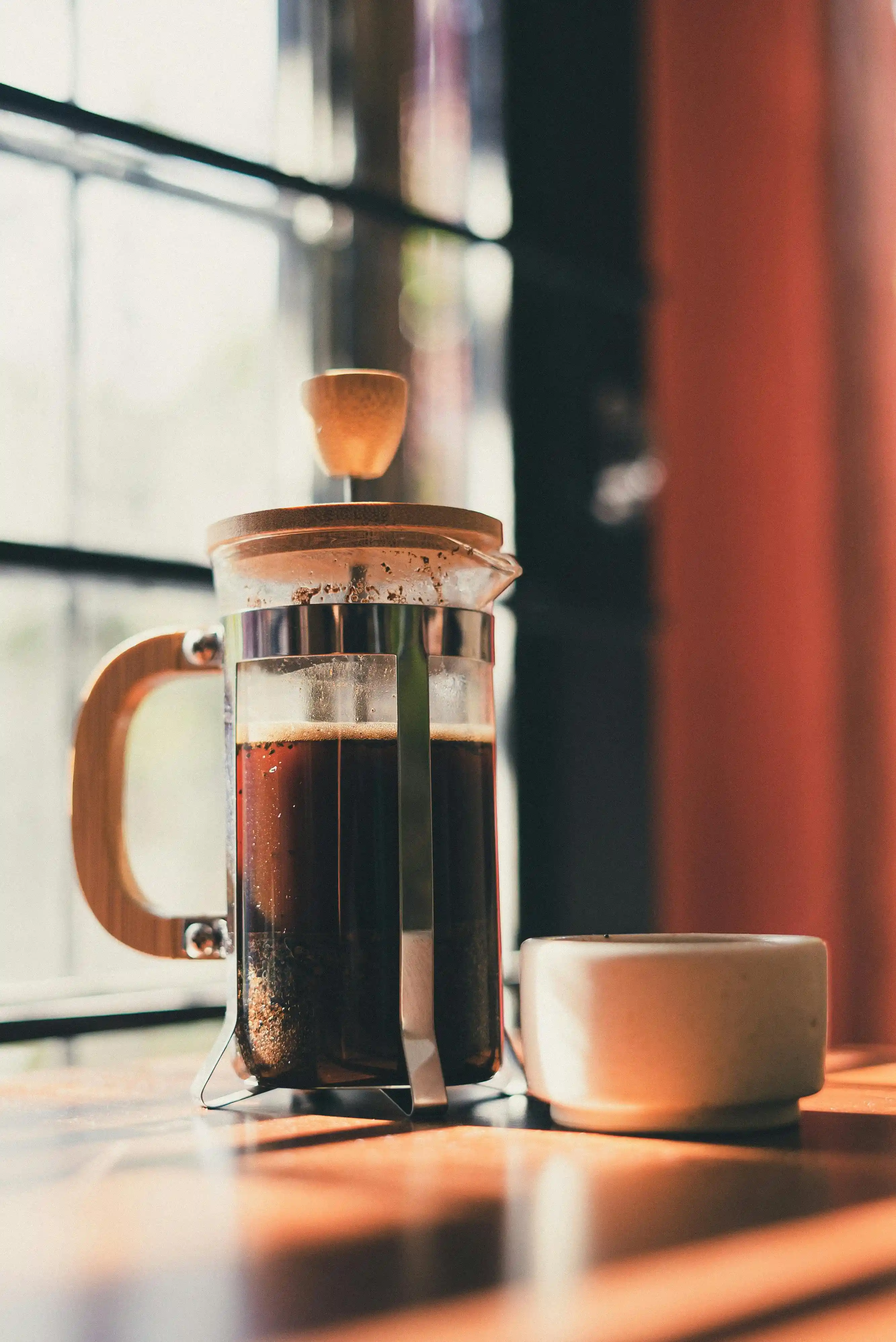 A glass french press filled with dark coffee sits on a sunlit wooden table near a window. Warm light highlights the rich color of the brew, with a simple ceramic cup resting beside it. The scene feels calm and inviting, capturing a quiet moment made for slowing down and savoring.