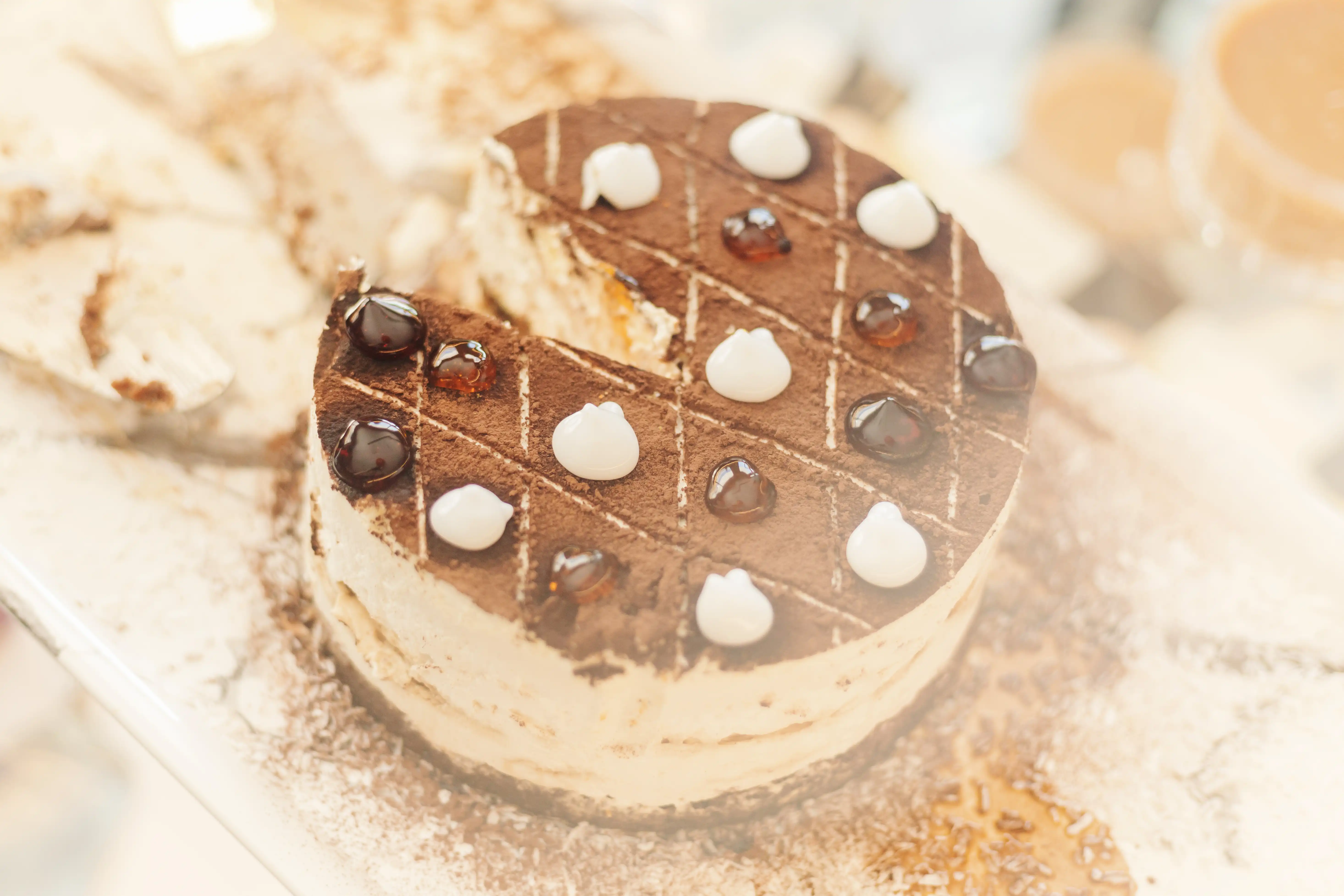 An appetizing ice cream cake is presented on a white platter, featuring a dusting of cocoa powder and a distinctive diamond-patterned top. The dessert, known in French as a gâteau à la crème glacée, is decorated with alternating dollops of white cream and dark chocolate glaze. One slice has been removed, revealing the creamy layers and rich chocolate base of this elegant treat.