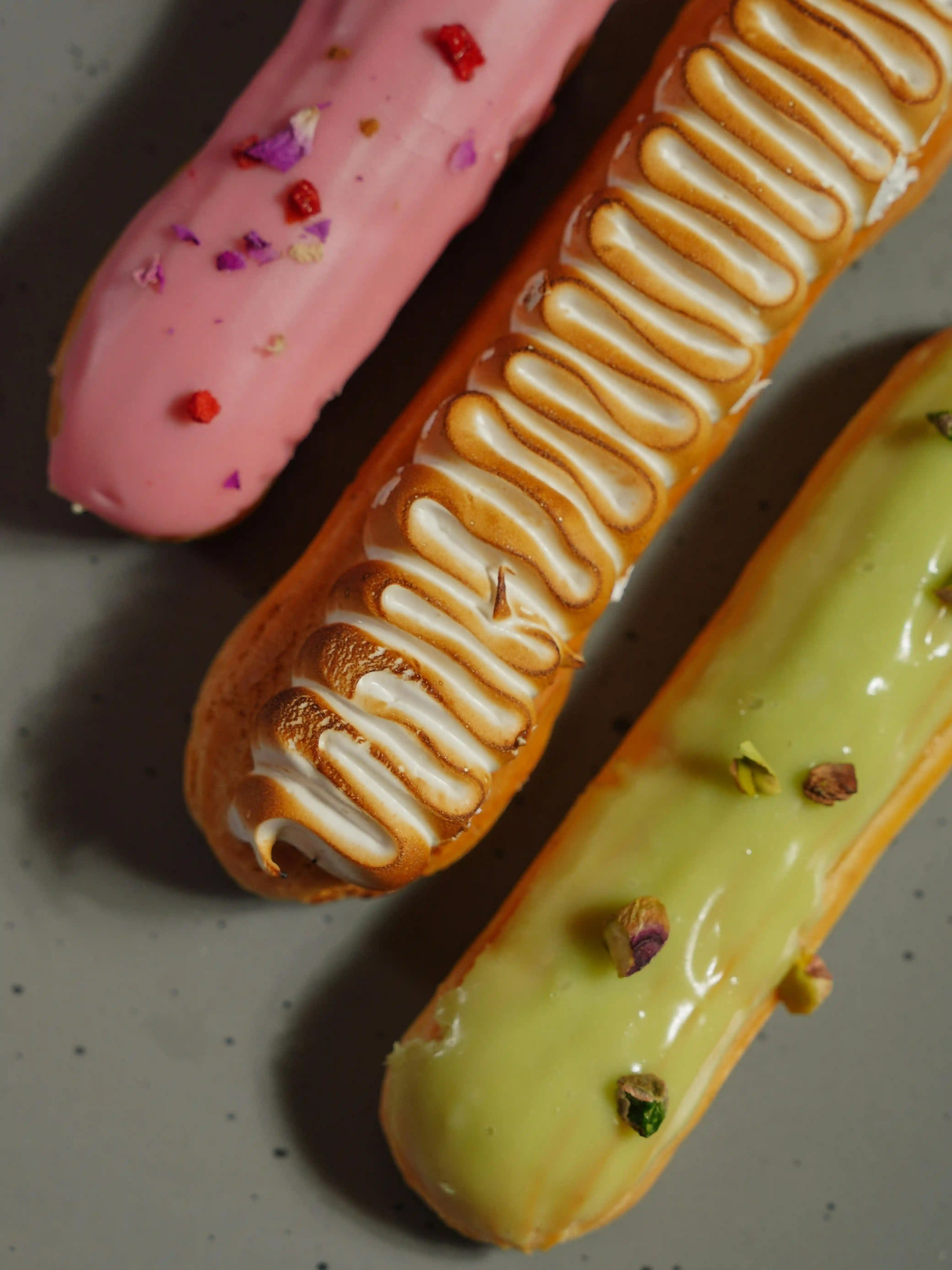 Assorted éclairs with pink glaze, toasted meringue piping, and pistachio icing rest on a speckled plate. For éclair shells, the unbleached flour vs bleached choice can affect how tender yet structured the pastry feels once baked.