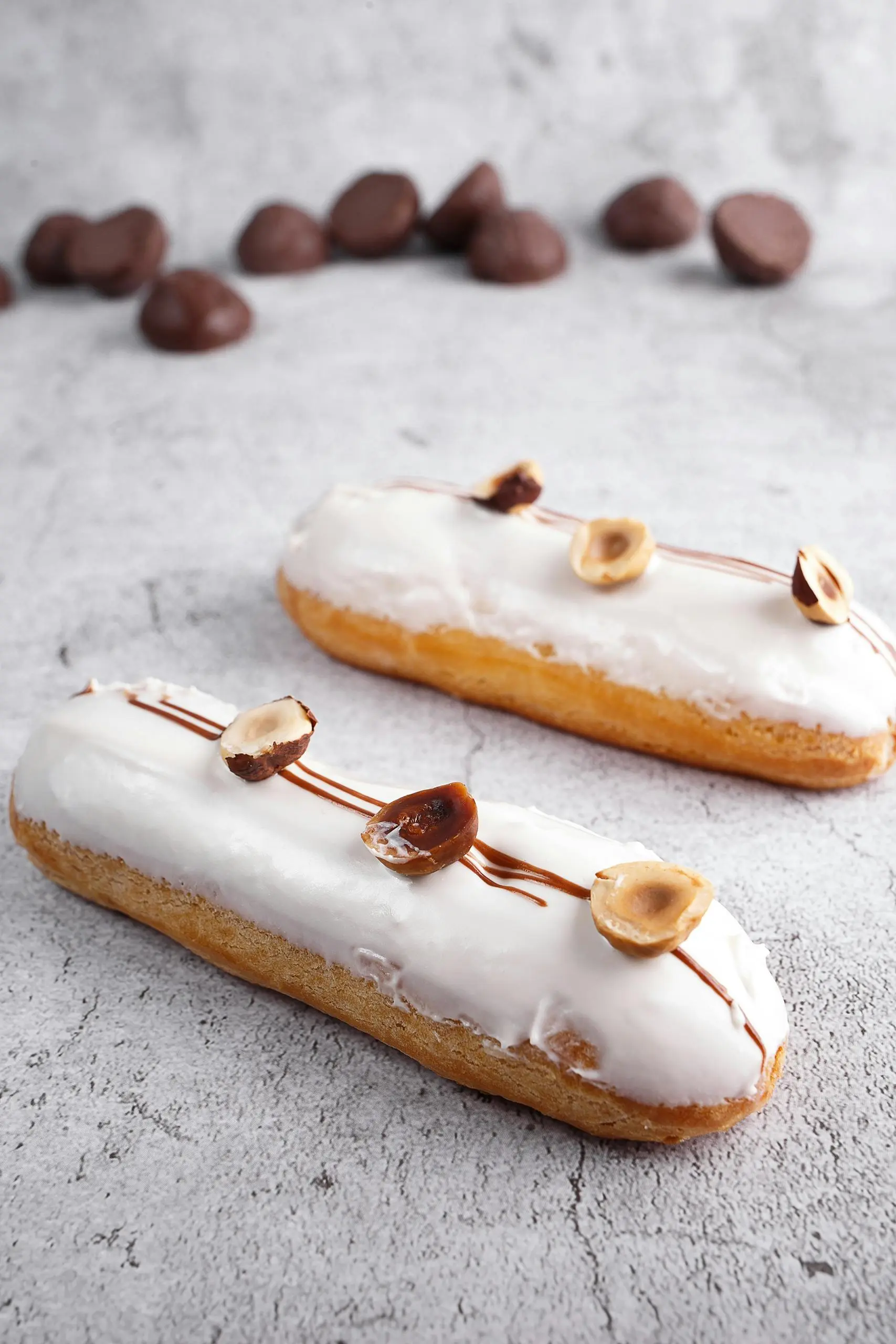 A pair of elegant éclairs is topped with a smooth, creamy vanilla glaze and garnished with roasted hazelnut halves. Thin caramel drizzles crisscross the topping, which is flavored with premium vanilla beans from Madagascar. The pastries are displayed on a neutral stone surface with dark chocolate pieces scattered in the background.